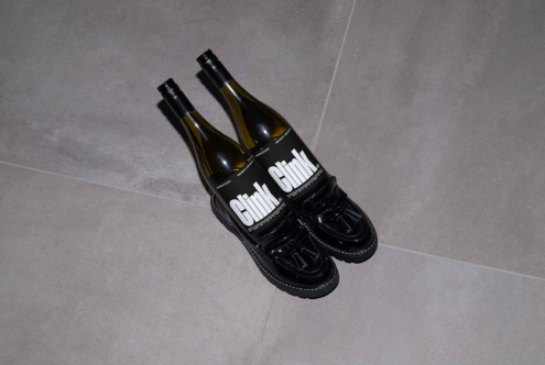 Two wine bottles creatively set as shoes with black glossy loafer shoes on gray tiled floor. Clink branding visible. Unusual product design mockup.