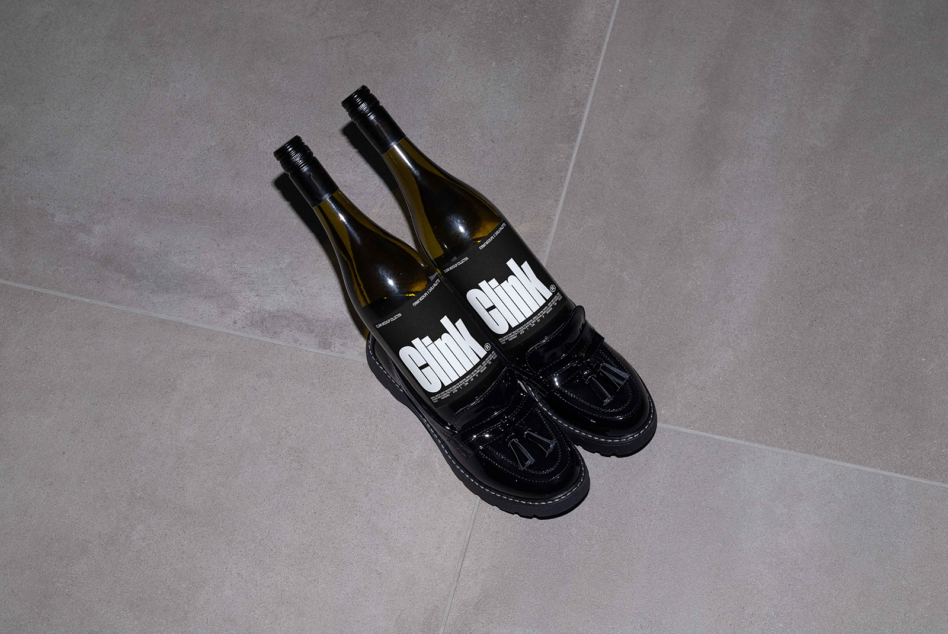 Two wine bottles creatively set as shoes with black glossy loafer shoes on gray tiled floor. Clink branding visible. Unusual product design mockup.