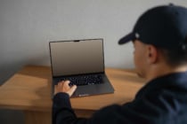 Person wearing black cap working on laptop at wooden desk