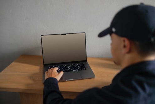 Man in cap using laptop on wooden desk with blank screen perfect for digital mockup designs and templates ideal for designers and creatives