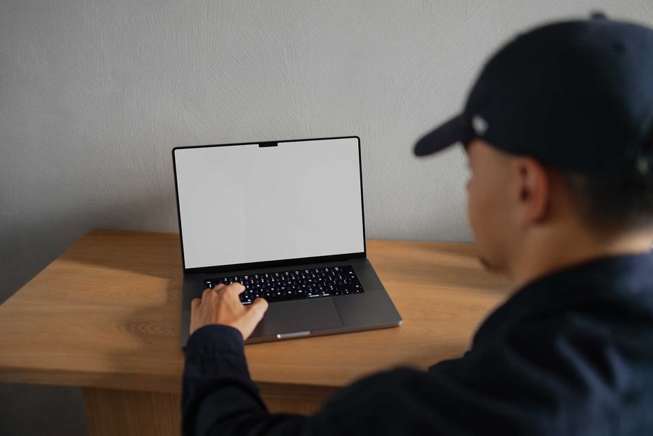 Person in black cap typing on laptop with blank screen at wooden desk