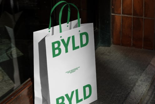 Shopping bag mockup with green BYLD logo, thick paper, sturdy handles, shadowed urban setting, ideal for branding, packaging design, graphic resources.