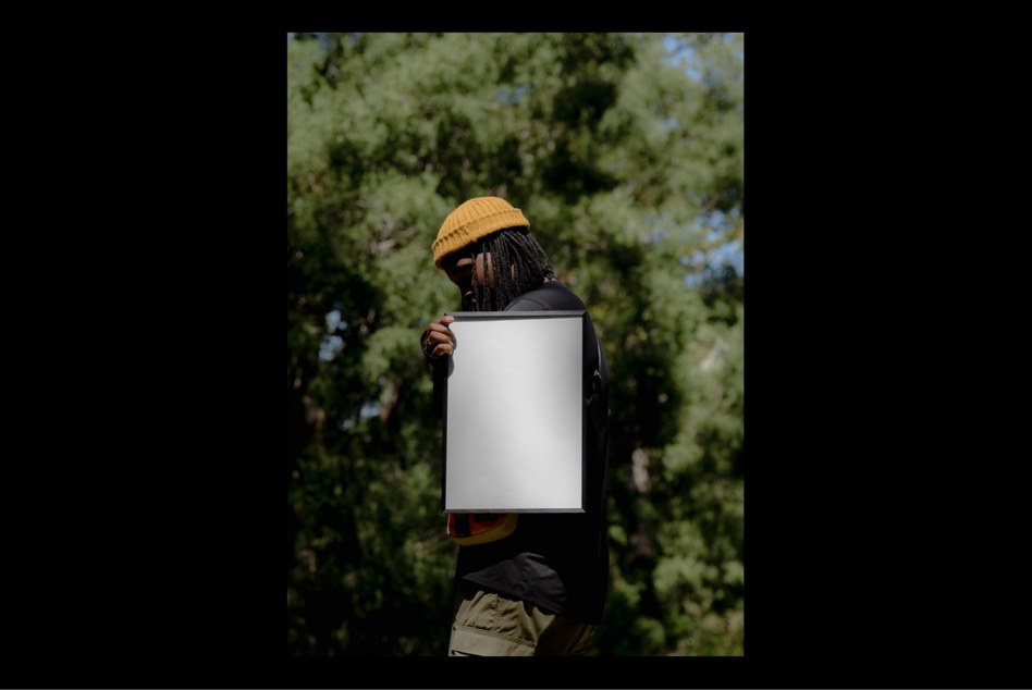 Man in a yellow hat holding blank poster mockup for design projects in an outdoor setting with green trees; ideal for showcasing graphics or templates.