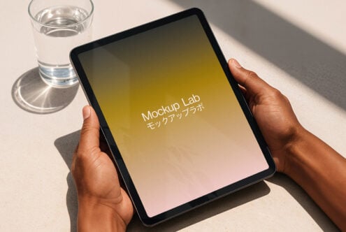 Tablet mockup held by hands on a table with a glass of water. Ideal for designers showcasing apps. Keywords: tablet mockup, digital assets, design.