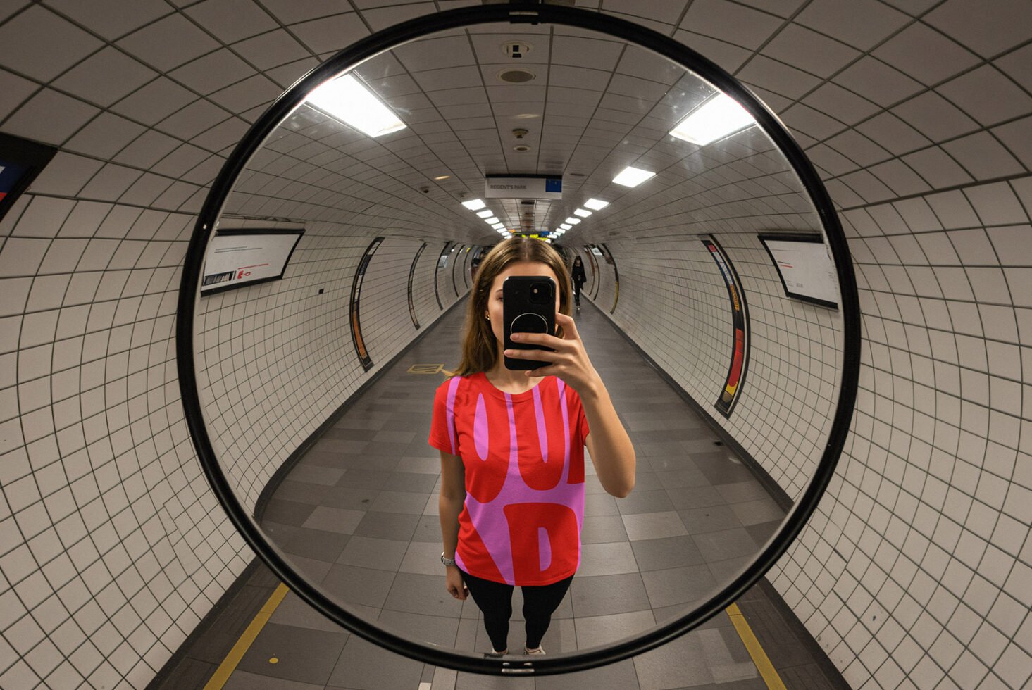 Woman in subway wearing bold graphic tee captures reflection in convex mirror. Mockup ideal for designers showcasing urban-inspired apparel graphics.