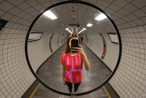 Woman in subway wearing bold graphic tee captures reflection in convex mirror. Mockup ideal for designers showcasing urban-inspired apparel graphics.