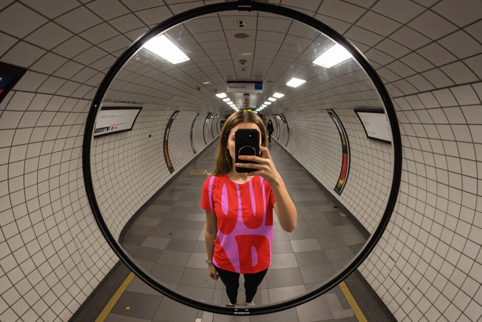 Person taking phone mirror selfie in tunnel wearing red magenta graphic tee