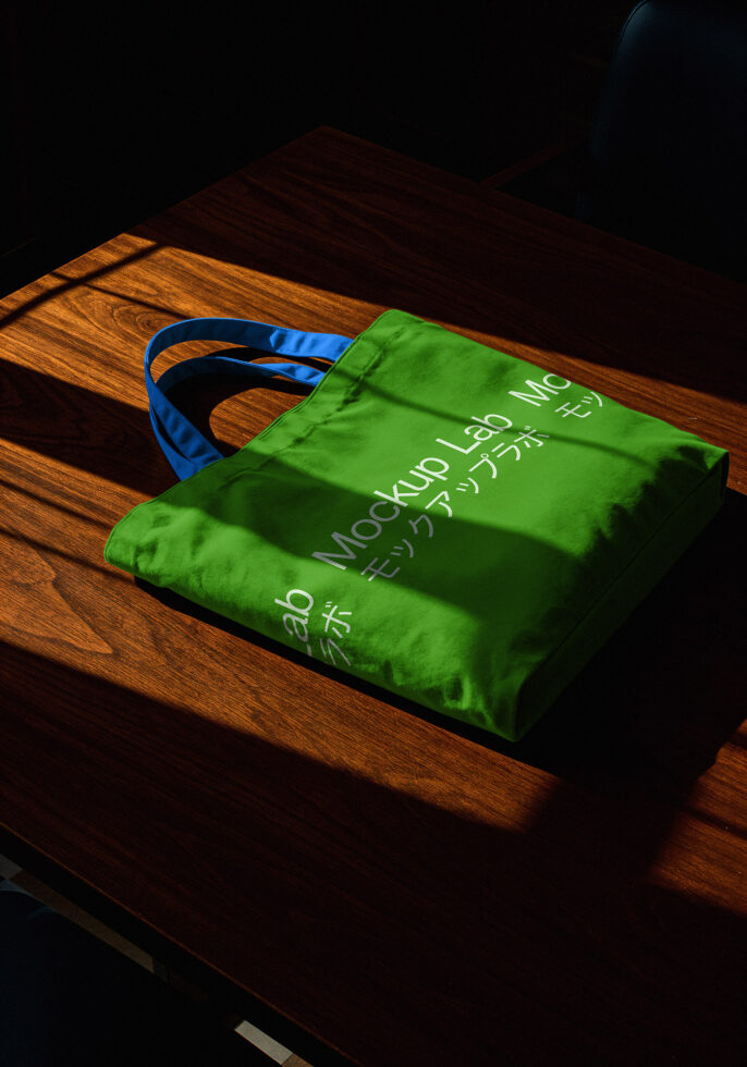 Green tote bag mockup with blue handles on wooden table. Perfect for showcasing branding designs for portfolios. Keywords: mockup design branding tote.