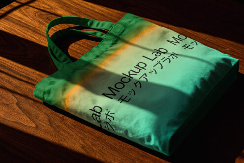 Green tote bag mockup with shadow overlay on wooden surface featuring Japanese text. Ideal for showcasing branding designs. Perfect for graphic designers.