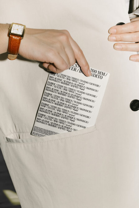 Close-up of hand with watch inserting printed typography fabric label into beige coat pocket. Suitable for mockups and graphic design projects.