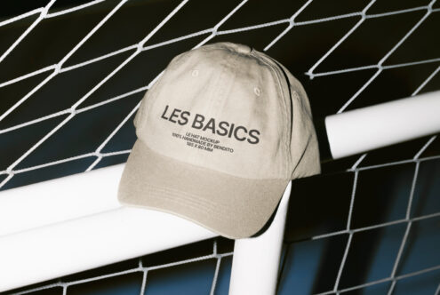Beige cap hat mockup displayed against a wire fence. Features text saying Les Basics. Perfect for showcasing hat designs and branding concepts for designers.