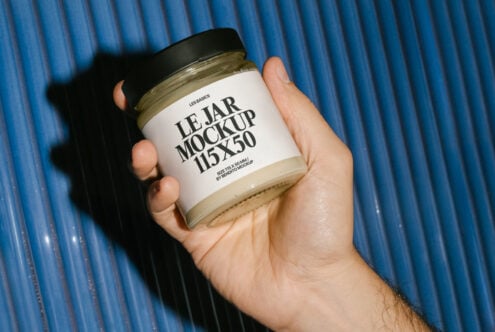 Hand holding a jar with label mockup against blue background. Ideal for designers seeking packaging mockups, branding, product design, and graphics.