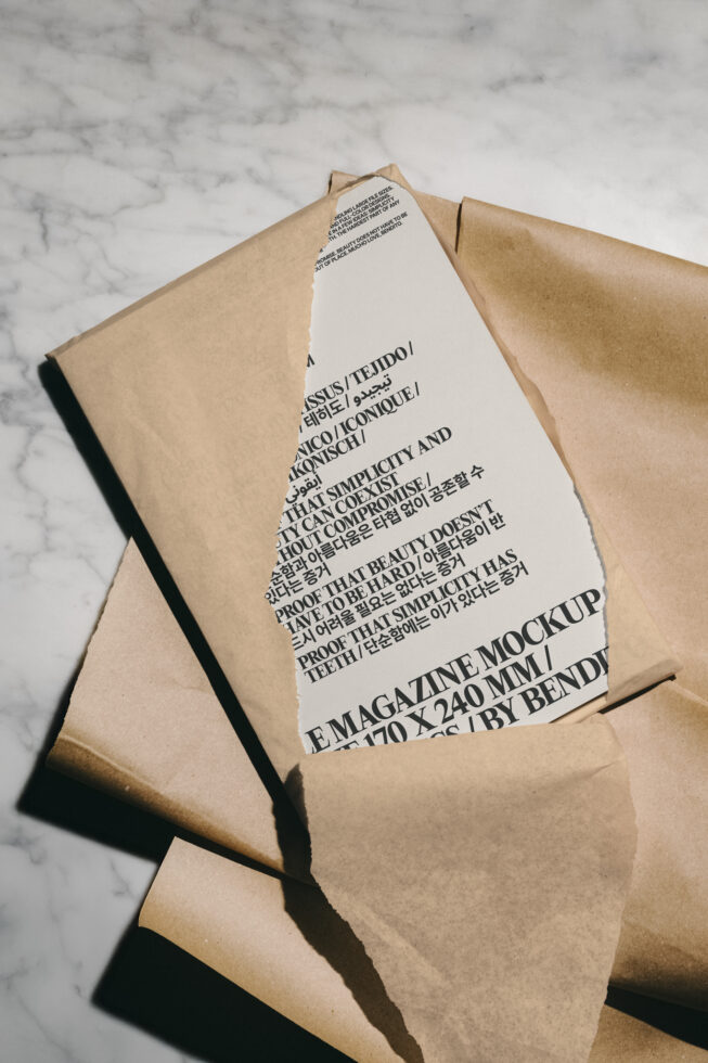 Magazine mockup partially revealed from torn brown paper on marble surface, showcasing minimalist design with bold typography. Ideal for designers' templates.