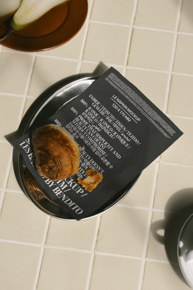 Napkin mockup on a tiled surface with a croissant on a metal tray. Perfect for showcasing typography design, product mockups, and template presentations.