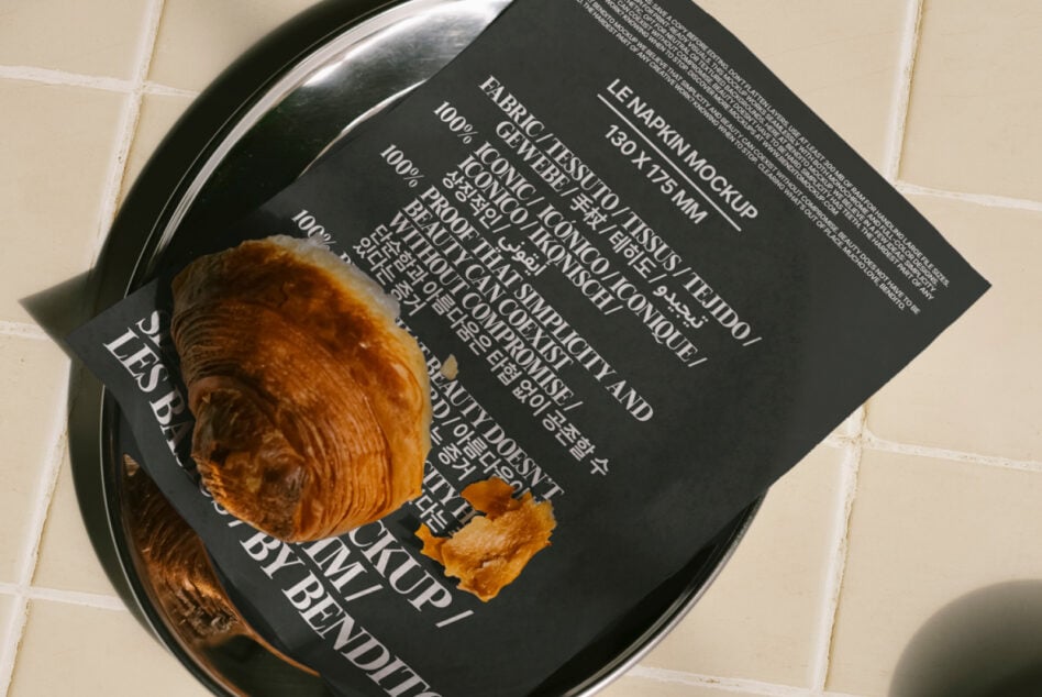 Napkin mockup featuring a croissant on a dark print napkin set on a shiny plate on a tiled background. Keywords: mockup, napkin, croissant, design asset.
