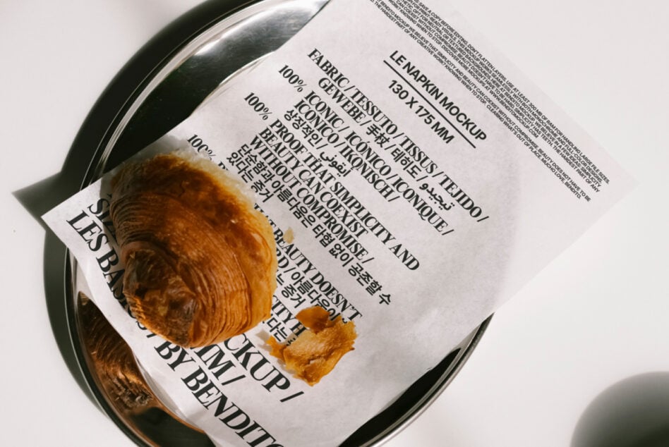 Napkin mockup featuring text in multiple languages on a shiny tray with a croissant. Perfect for design projects, templates, and print presentations.
