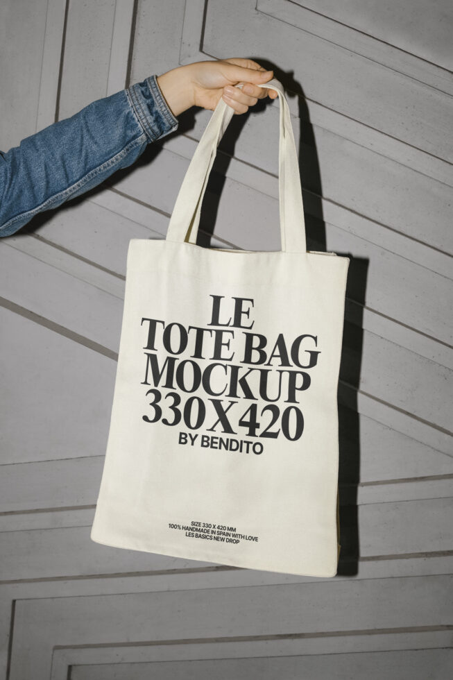 Tote bag mockup held by a hand with denim sleeve against a textured wall. Perfect for showcasing design templates and branding ideas. Eco-friendly material.