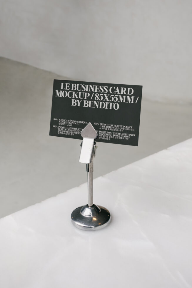 Black business card mockup held by desk clip stand with multilingual text on white surface