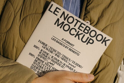 Notebook mockup held by a person in a green jacket, featuring multilingual text on the cover. Ideal for designers seeking sleek, editable mockup templates.