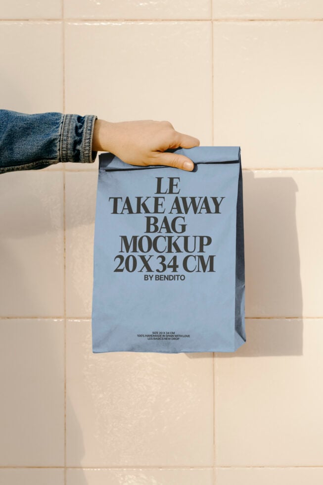 Hand holding blue takeaway bag mockup 20x34cm by Bendito against tiled background. Ideal for designers looking for packaging templates and graphics.
