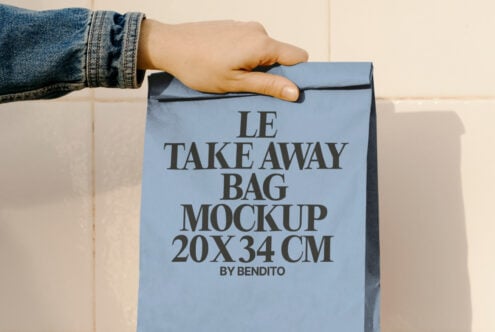 Blue paper take away bag mockup held by hand. Text reads LE TAKE AWAY BAG MOCKUP 20x34 CM. Ideal for packaging design, product showcase, branding.