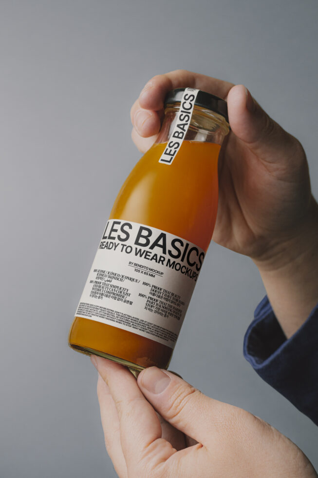 Mockup of hands holding a glass juice bottle with LES BASICS branding. Ideal for designers needing packaging mockups for presentations or promotional graphics.