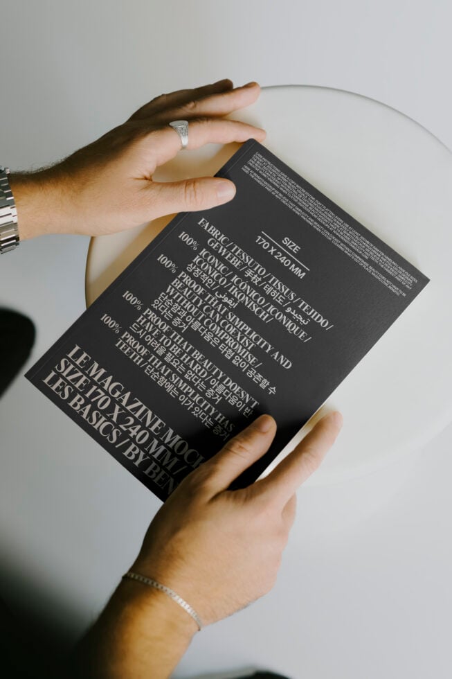 Magazine mockup held by hands wearing jewelry on a minimal background. Black cover with multilingual text. Ideal for designers focused on typography.