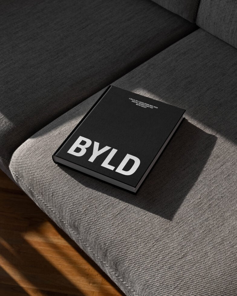 Mockup of a hardcover book titled BYLD resting on a gray sofa in natural light. Ideal for showcasing book cover designs or graphic projects.