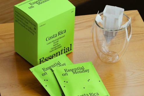 Bright green coffee packaging with text Costa Rica and Essential on Monday next to a glass mug with coffee filter on wooden table. Perfect for branding mockups.