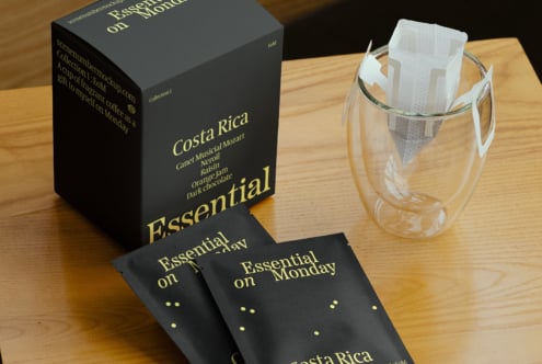 Coffee packaging mockup featuring a minimalist design with Costa Rica text in yellow on black, accompanied by a glass mug displaying a drip bag. Ideal for designers.