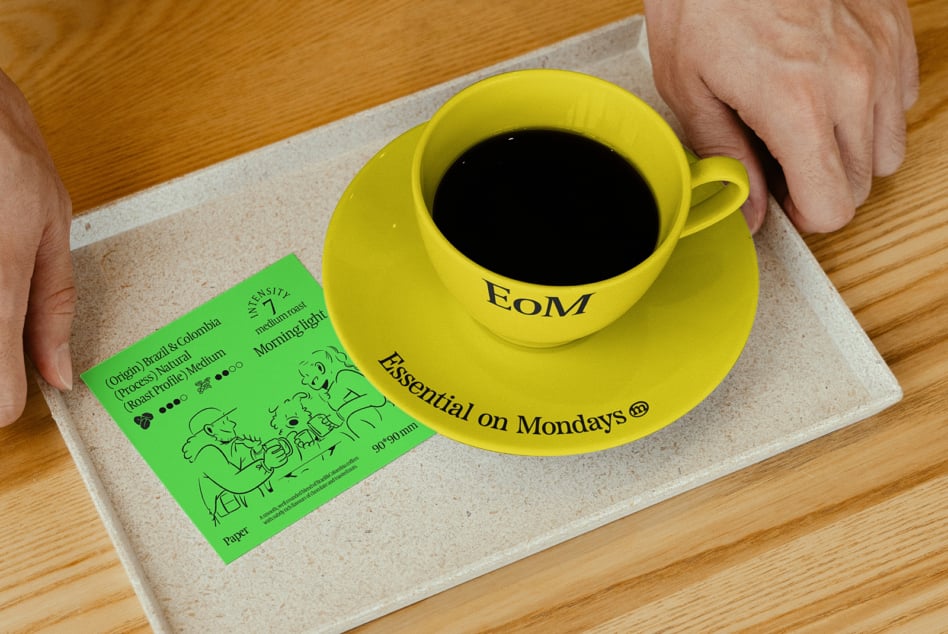 Yellow coffee cup with text Essential on Mondays on saucer placed on tray beside a green card with coffee details perfect for branding mockups designers