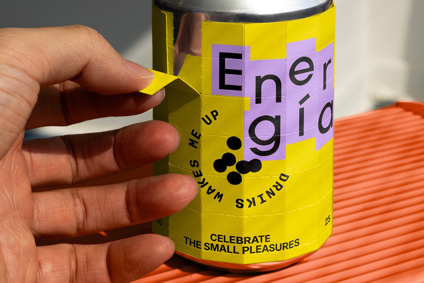 Hand peeling yellow and purple sticker on can with bold typography Celebrate the Small Pleasures; keywords include Mockup, Packaging Design, Label Graphics