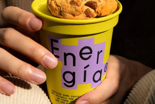 Person holding a vibrant yellow coffee cup with modern energy logo and cookie on lid suitable for branding mockup design lifestyle packaging concept.