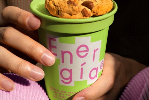Green cup with pink text reading Energia held by hands. Topped with a cookie. Ideal for product mockups branding design or digital assets.