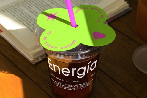 Iced coffee with vibrant green lid and pink straw on a wooden table beside an open book. Keywords: mockup, graphic design, template, vibrant, energy.