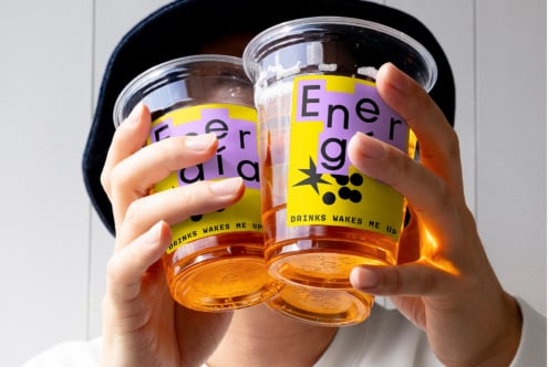 Close-up image of hands holding two clear plastic cups with vibrant purple and yellow labels reading Energaia. Perfect for branding mockup or design template.