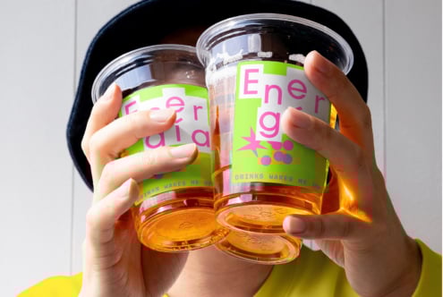 Close-up of hands holding two plastic cups with colorful graphic labels featuring energy drink branding. Ideal for mockups or design presentations.