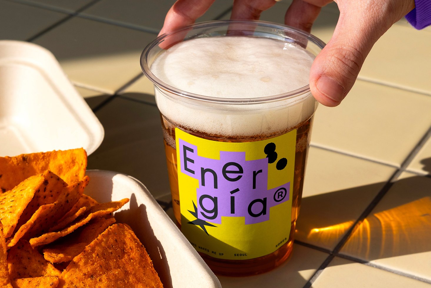Close-up of a hand holding a cup of beer with colorful branded label, next to a tray of nacho chips, ideal for mockups or food branding templates.