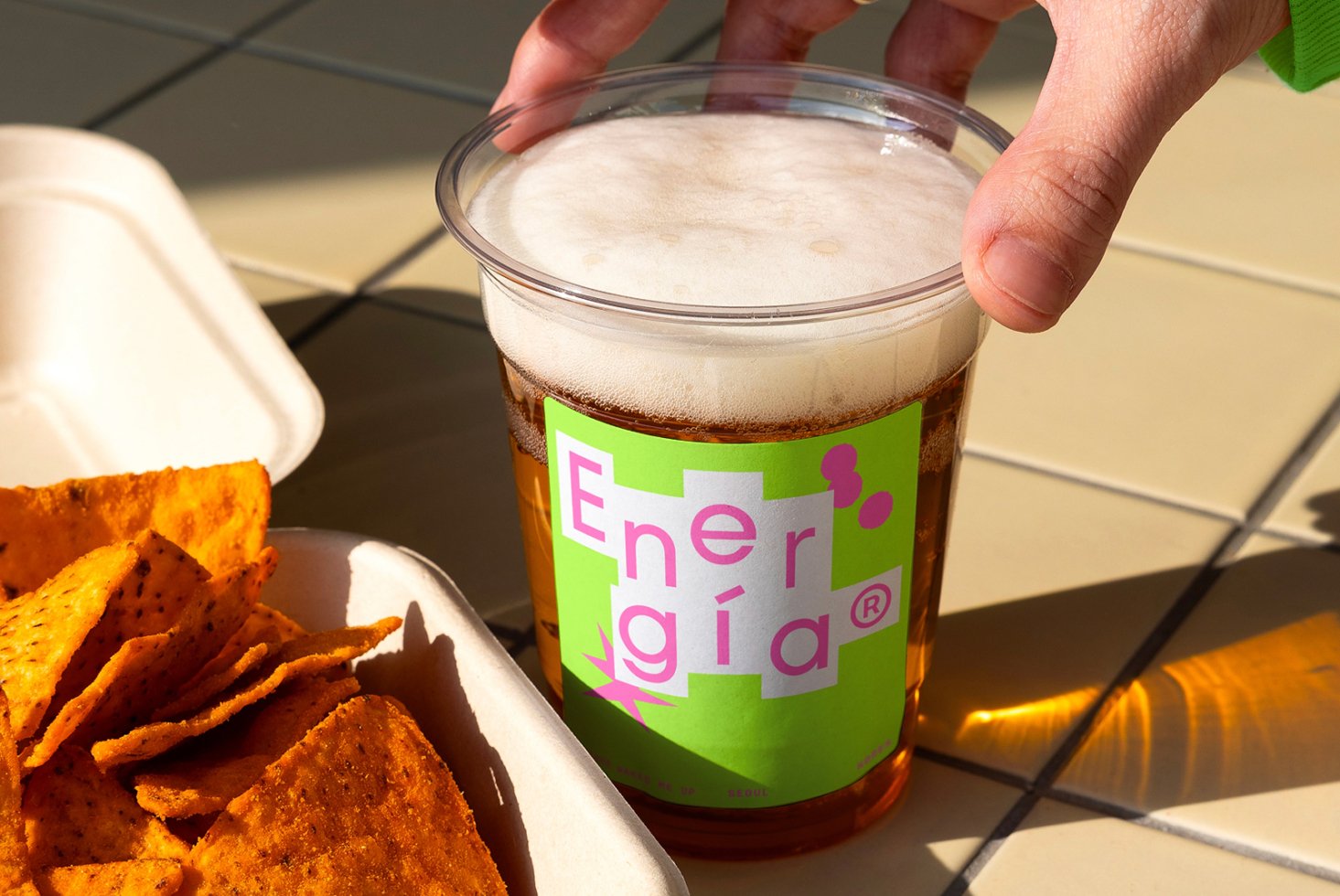Hand holding cup with green label design next to tray of chips on tiled surface. Perfect for mockups or packaging templates. Bright outdoor setting.