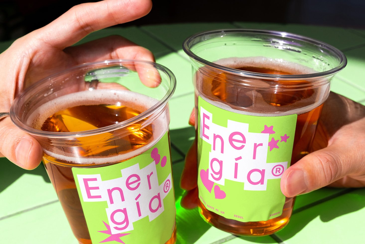 Hands holding cups with vibrant green and pink labels saying Energia. Ideal for beverage branding mockups. Bright design and outdoor setting.