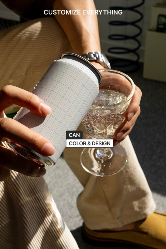 Mockup image of a hand holding a customizable grid-patterned can beside a glass of sparkling water. Perfect for branding, packaging design projects.