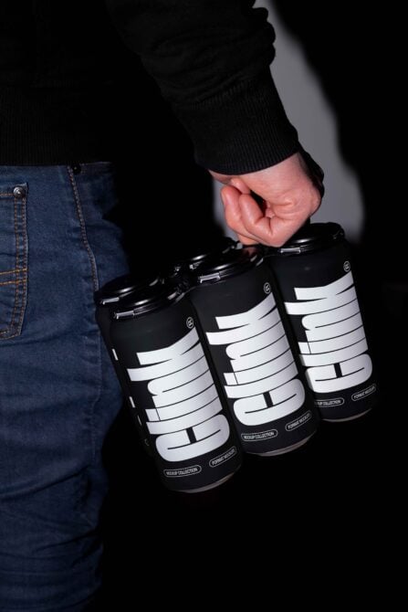 Hand holds black drinks cans with bold text in modern font. Jeans visible in background. Ideal for branding mockup, packaging design, marketing visuals.