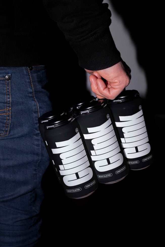 Hand holds black drinks cans with bold text in modern font. Jeans visible in background. Ideal for branding mockup, packaging design, marketing visuals.