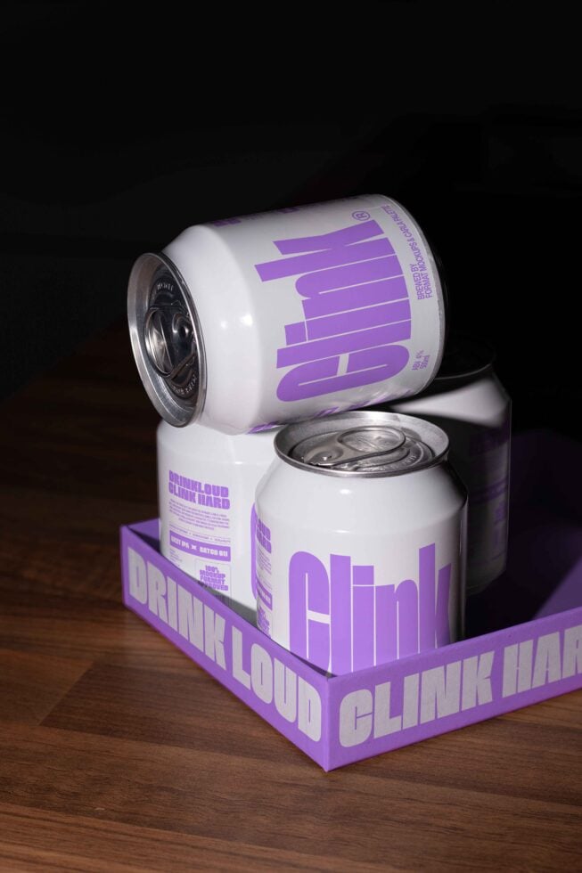 Mockup of three white soda cans with purple Clink branding on a wooden surface. Ideal for beverage design presentations, packaging mockups, template resources.
