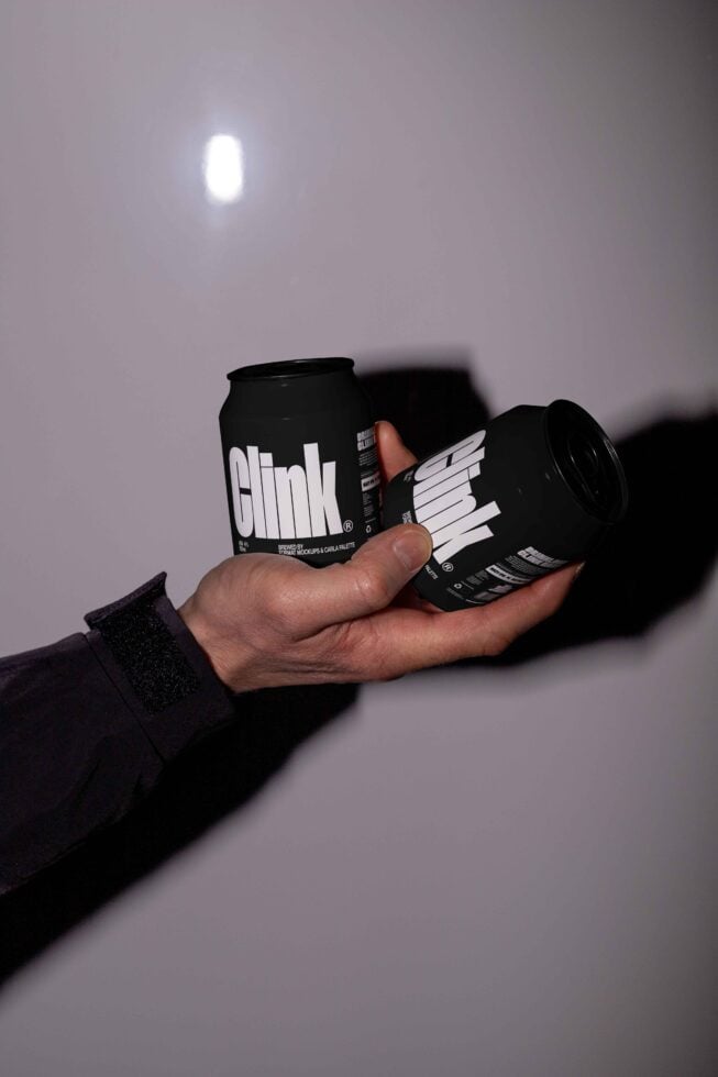 Hand holding two black beverage cans with bold text design on a reflective surface. Perfect for branding mockup templates and graphic design projects.
