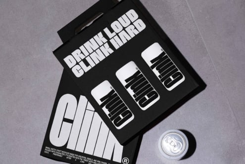 Mockup of a branded black cardboard carrier with bold typography design featuring the words Drink Loud Clink Hard next to a can on a gray surface.