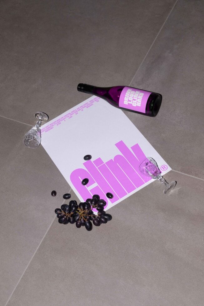 Wine mockup featuring a purple bottle on a gray surface with scattered grapes and glasses. Ideal for designers seeking elegant product presentation.