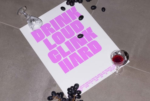 Poster mockup with bold pink text DRINK LOUD CLINK HARD surrounded by scattered grapes and crystal glasses with red liquid on gray background.