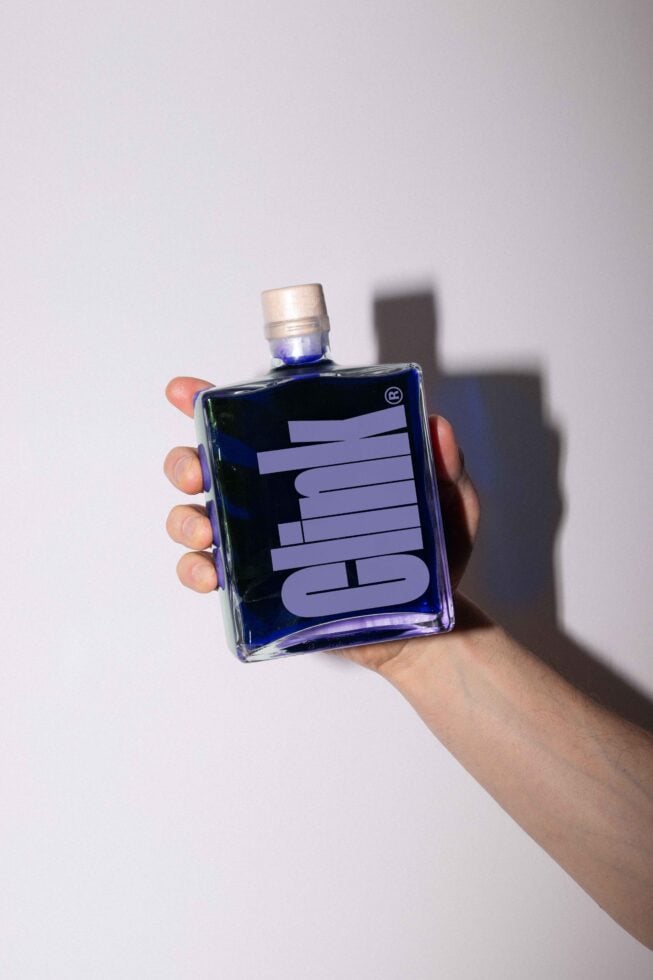 Hand holding a transparent square glass bottle with bold branding clink label. Perfect mockup for designers. Product packaging graphic asset.