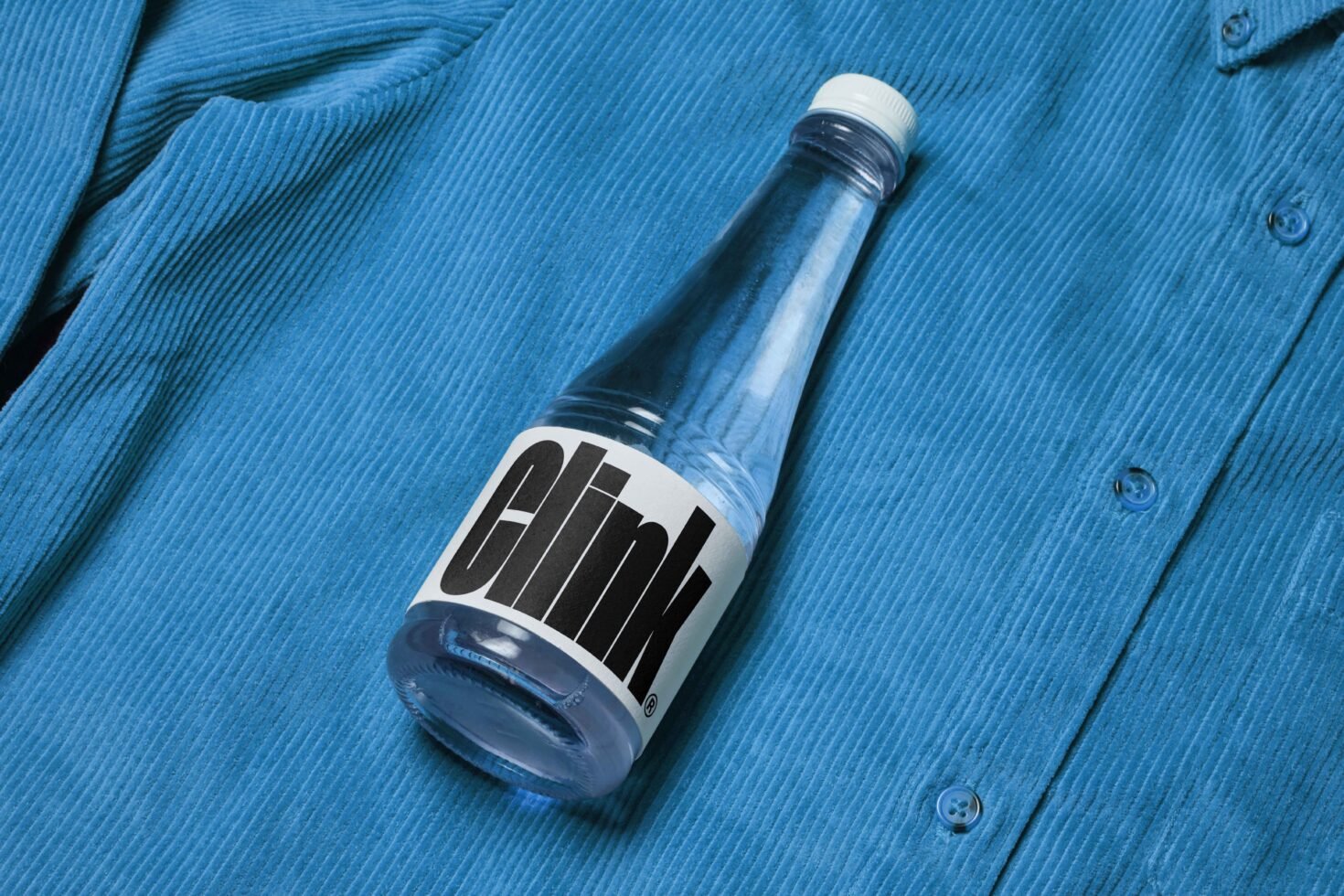Clear glass bottle with bold black typography label on blue textured fabric background. Ideal for product mockup, branding, packaging design.